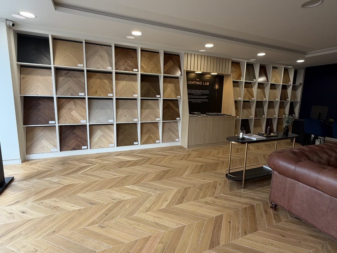 Wide view of the Hampton Hill Flooring showroom interior showing chevron oak flooring, display shelving for flooring samples, Chesterfield sofa seating area and the Lighting Lab feature wall